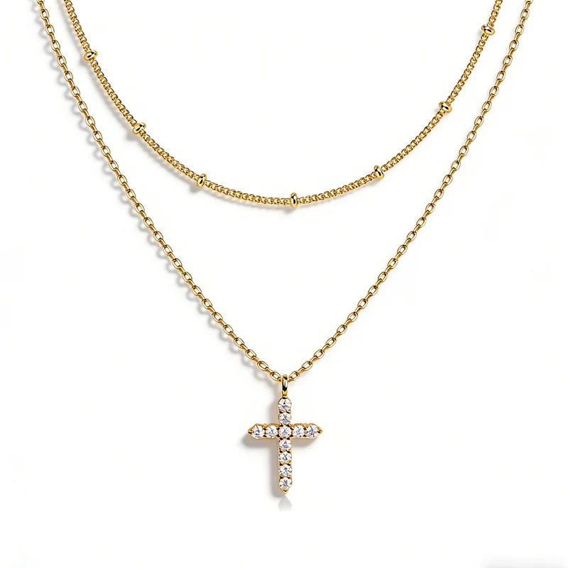 A double-layered gold necklace featuring a delicate chain and a longer beaded chain with a small cross pendant adorned with clear crystals, set against a plain white background.