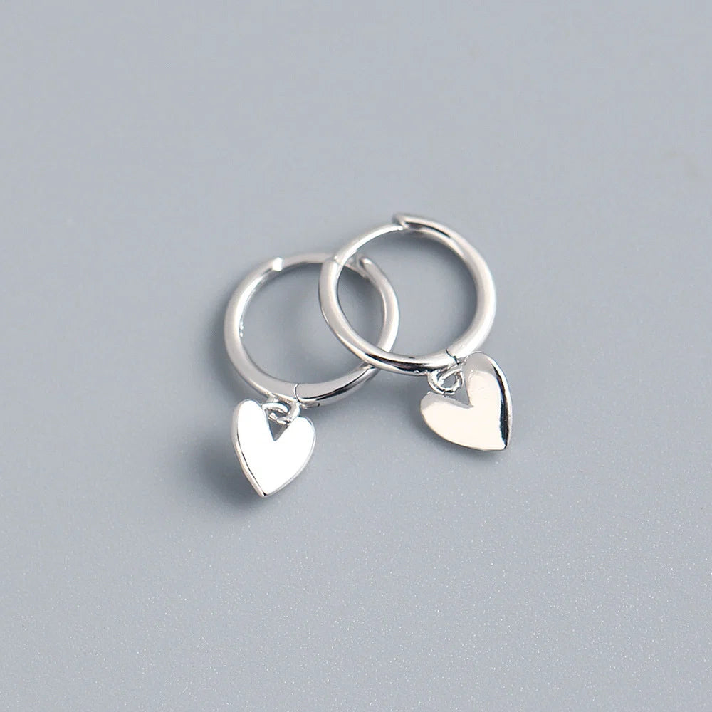 Two small silver hoop earrings with dangling heart charms, photographed on a smooth light gray surface.