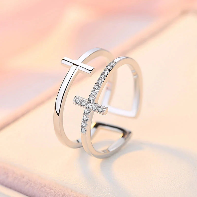 A silver open-shank ring featuring two crossed designs: one smooth polished cross and one adorned with small clear stones, displayed on a soft pink textured surface.