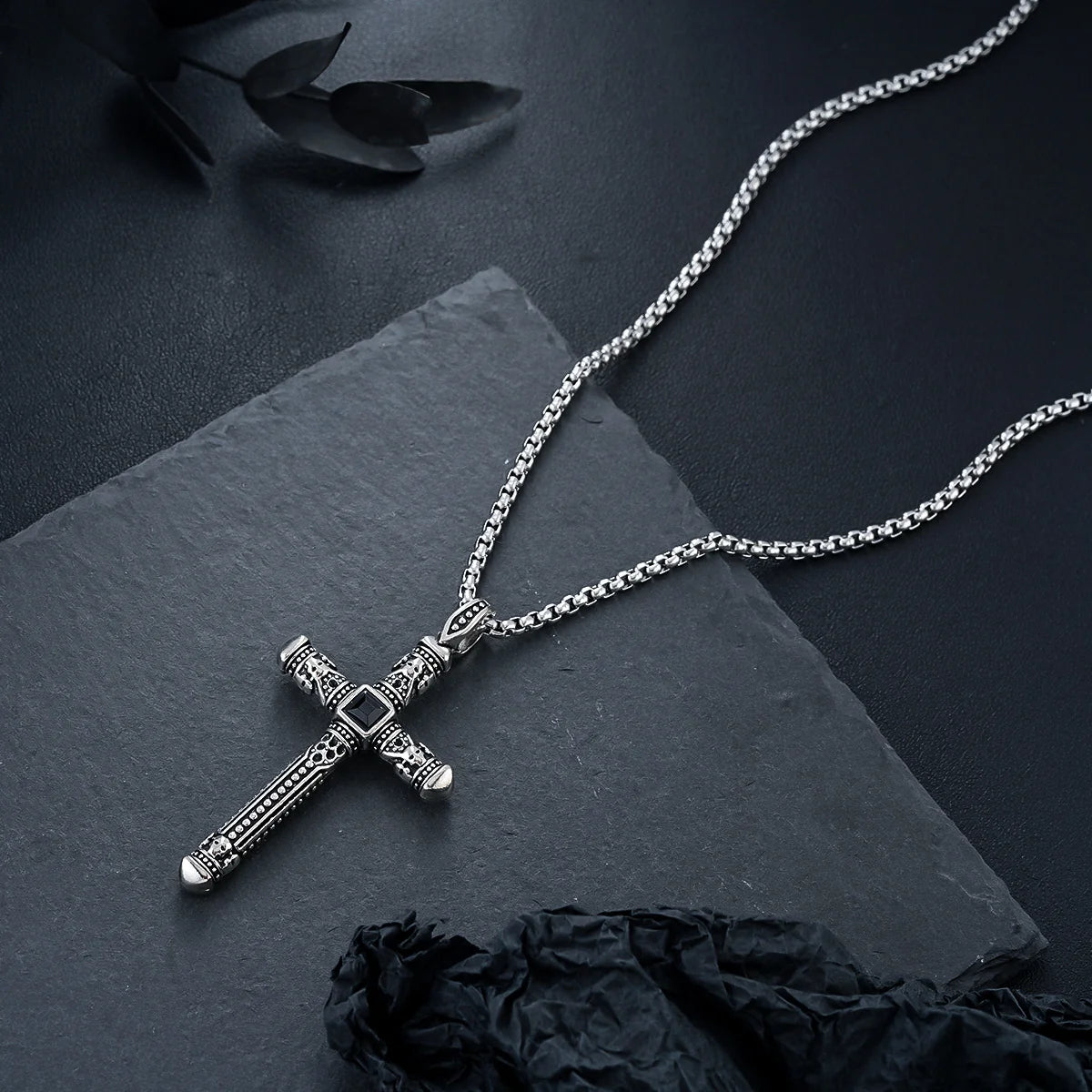 A silver-toned gothic cross pendant necklace with an ornate textured design and a central black square accent, displayed on a dark slate surface with black fabric and dried leaves.