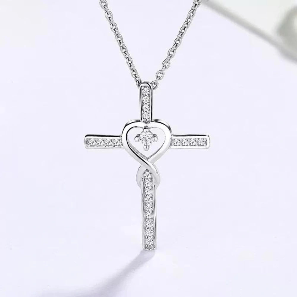 A silver cross pendant necklace adorned with small clear crystals, featuring a central heart-shaped crystal, hanging from a delicate silver chain against a clean white background.