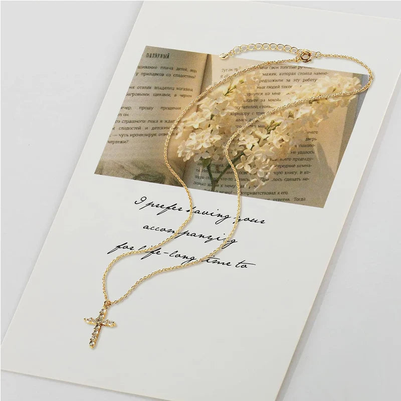 A delicate gold cross pendant necklace with a small clear crystal accent, displayed on a white card featuring a vintage book page with white flowers and cursive writing.