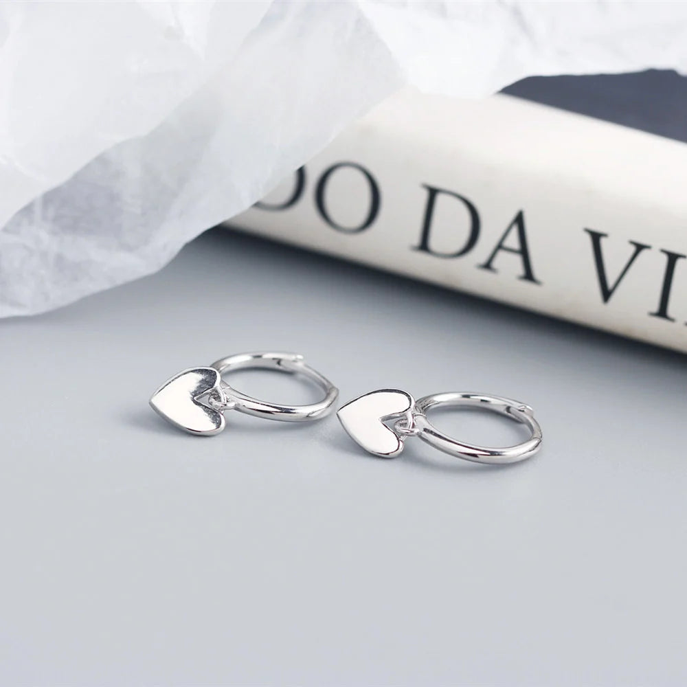 Silver heart-shaped hoop earrings on a light gray surface with a white fabric and magazine in the background.