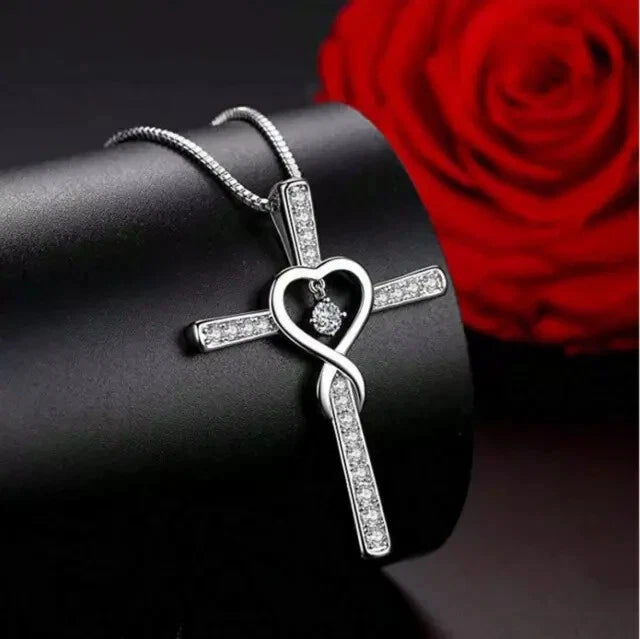 A silver cross pendant necklace with a heart design and clear stones, displayed on a black cylindrical stand with a red rose in the background.