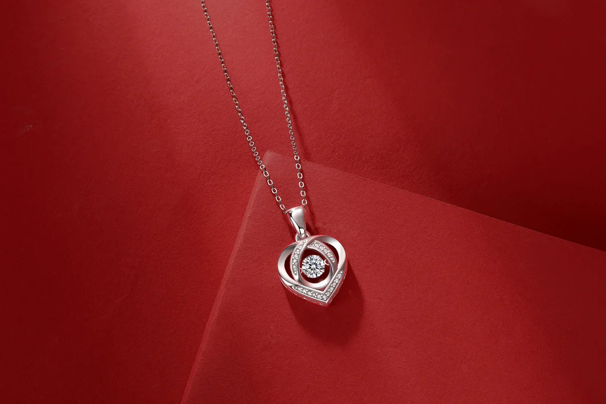 A silver heart-shaped pendant necklace with a central clear gemstone, displayed on a red textured background.