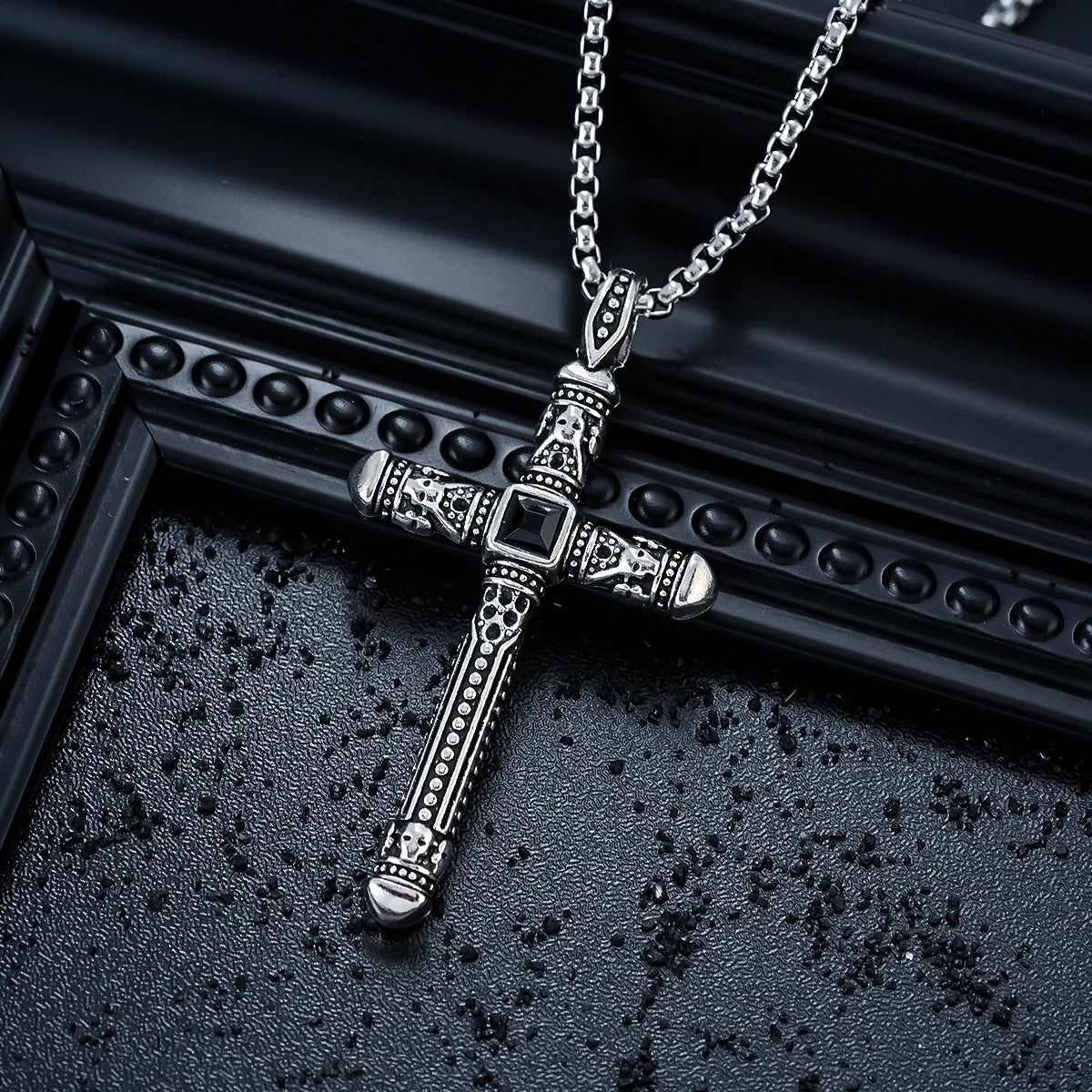 An ornate silver-toned cross pendant necklace with intricate filigree detailing and a central black square accent, resting on a textured black surface next to a black decorative frame.